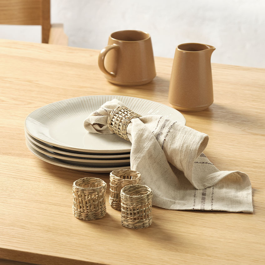 Beli krožniki z gubami na robu, vrč za mleko, prtički z tekstila in obročki za prtičke na jedilni mizi