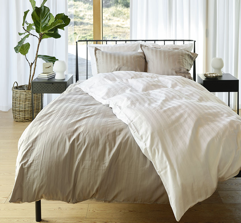 Spalnica s posteljnino v sivi in beli barvi z rahlimi črtami ter nočni omarici