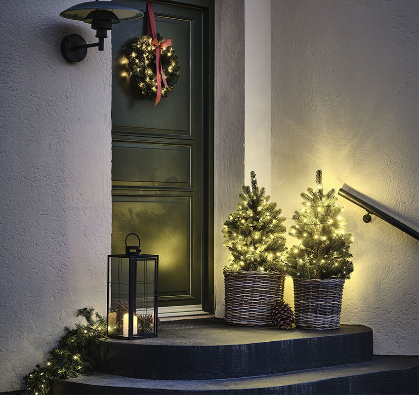 Dve božični drevesi pred vrati in adventni venček na vratih z rdečim trakom